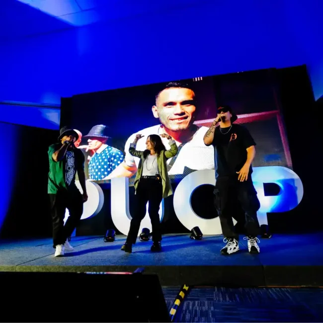 Trío de cantantes actuando en un escenario con luces azules frente a una gran pantalla